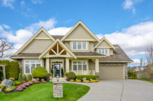Spacious two-story house with a gabled roof, front porch, and attached garage. Manicured lawn and shrubs in the foreground under a partly cloudy blue sky.